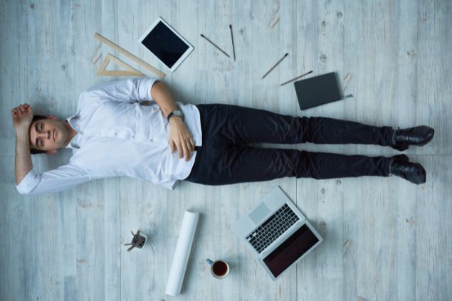 Imagem de um homem deitado no chão com computador, tablet e outros aparatos de escritório espalhados ao seu redor