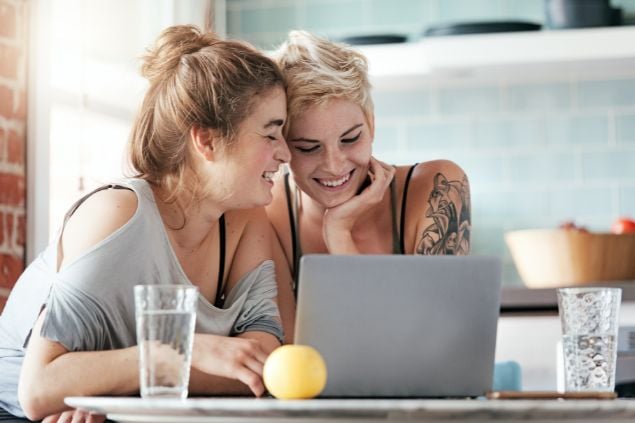 Imagem de um casal de mulheres olhando o notebook juntas e sorrindo