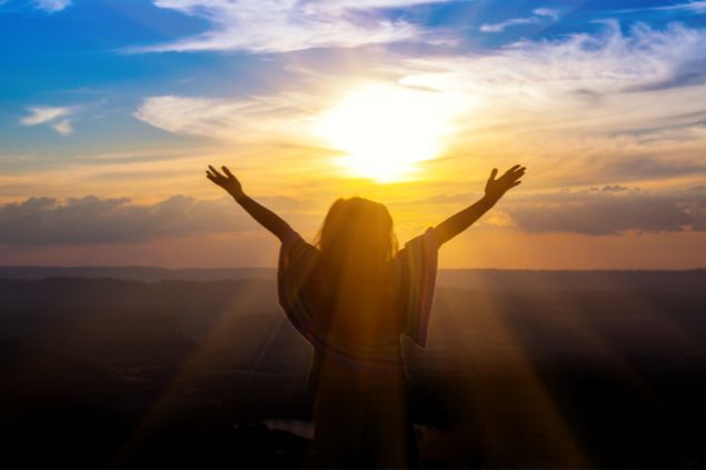 Imagem de uma pessoa com as mãos erguidas para o céu e aluz do pôr do sol a iluminando