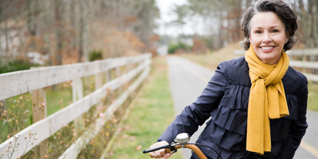Mulher andando de bicicleta em cenário bucólico. Ela está sorrindo e vestindo roupas quentes.