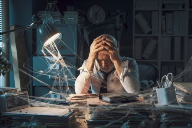 Imagem de um homem preocupado na frente de uma mesa de escritório cheia de teia de aranha, a imagem parece ilustrar uma crise financeira