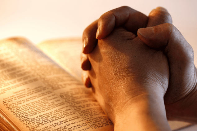 Mãos de homem entrelaçadas rezando sobre uma Bíblia. 