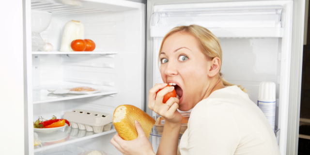 Mulher com semblante surpreso em frente a uma geladeira aberta. Ela está comendo.