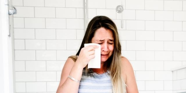 Mulher chora enquanto enxuga as próprias lágrimas com um lenço. Ela está em um banheiro. 